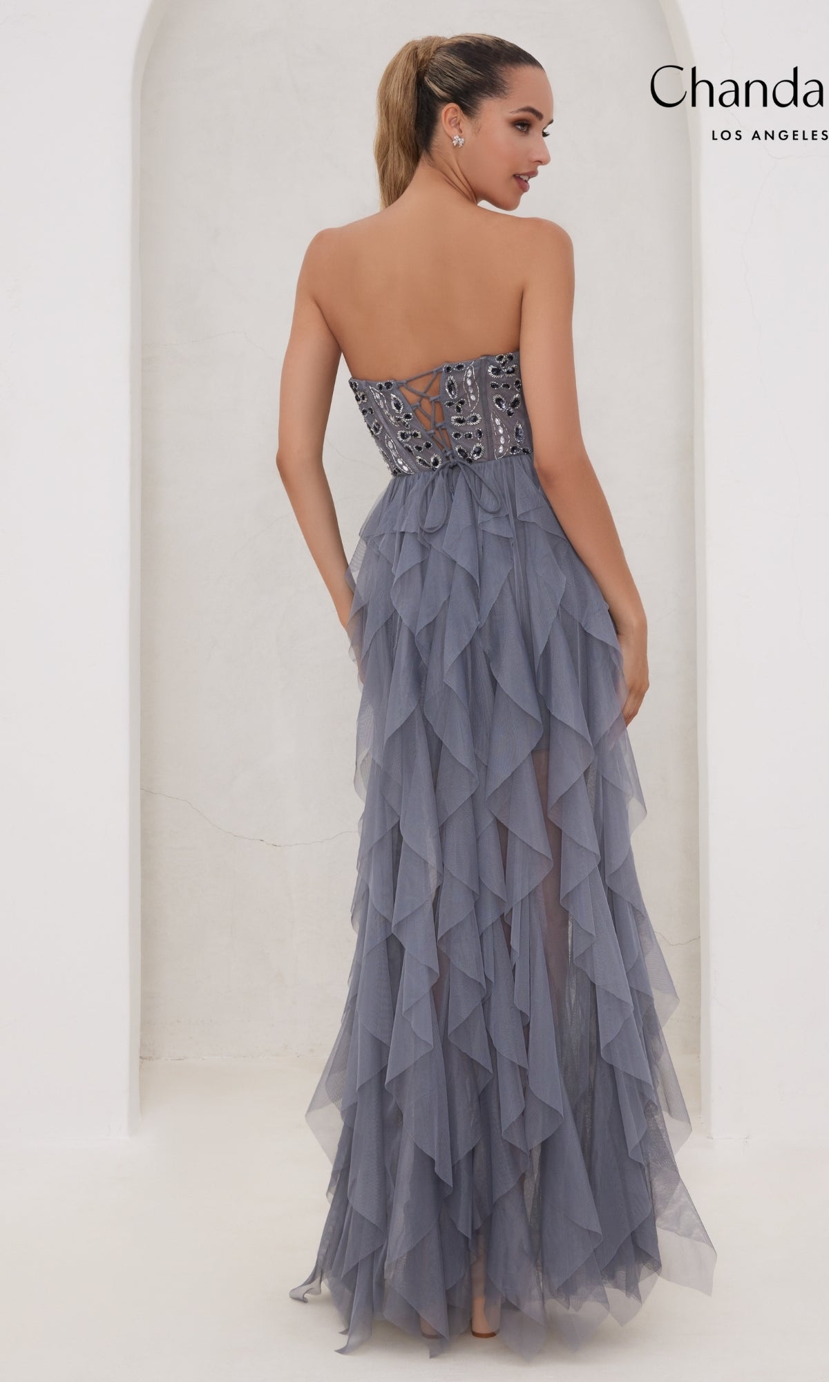 A woman in the Long Prom Dress 30151 by Chandalier, featuring a strapless beaded bodice and layered ruffle skirt, stands facing away with her hair in a ponytail.